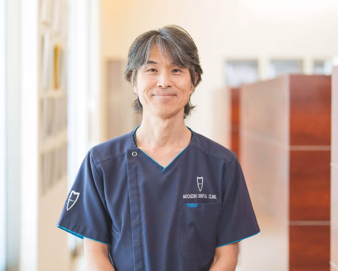 歯科医師が院内で笑顔を見せているポートレート写真。ユニフォームには「MOCHIZUKI DENTAL CLINIC」のロゴが入っている。