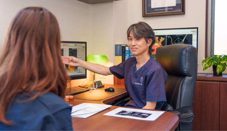 歯科医師がデスクで患者にモニターの画像を指しながら説明している様子。